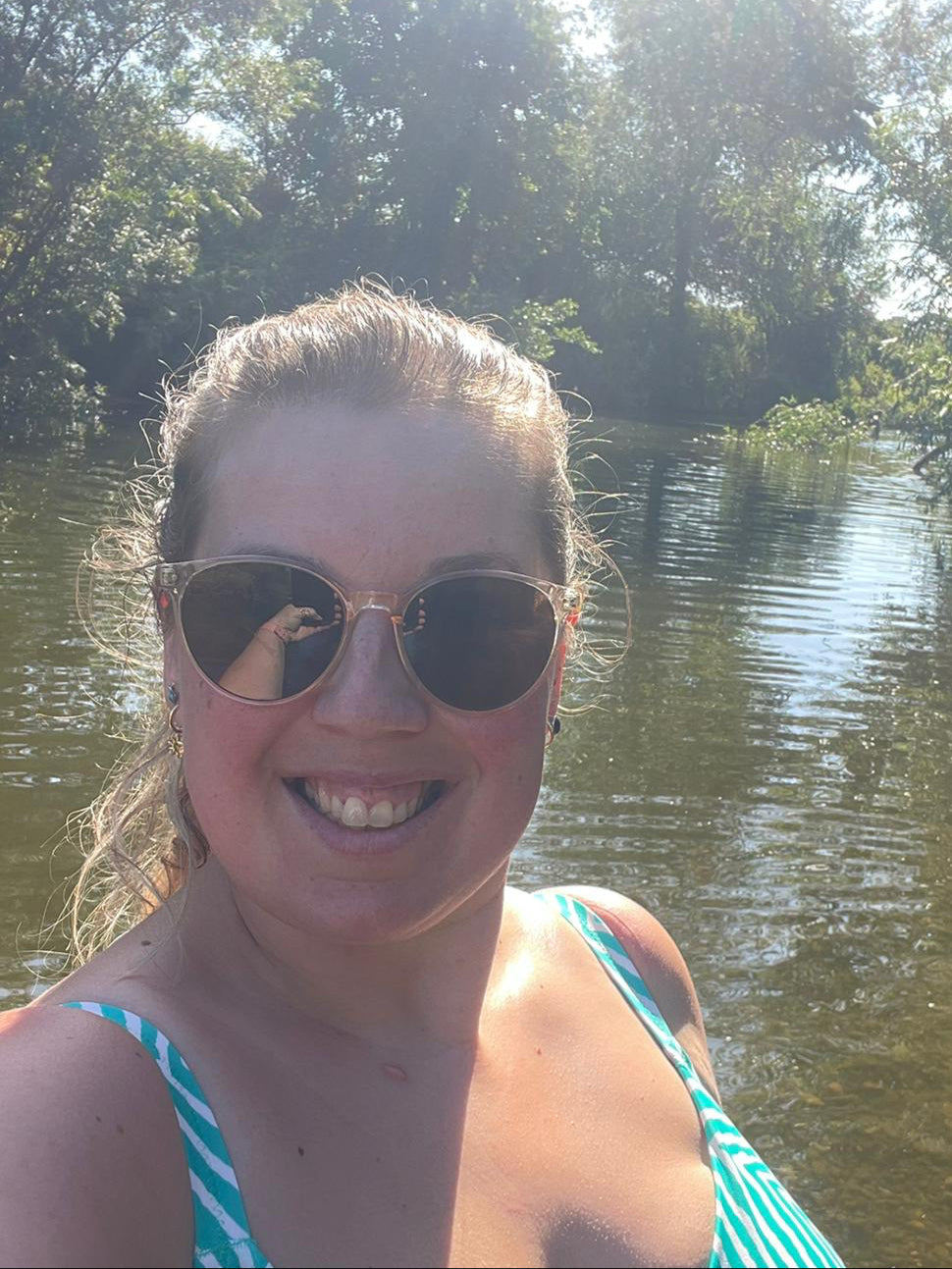 Woman wearing sunglasses and a striped swimsuit by a body of water with trees in the background